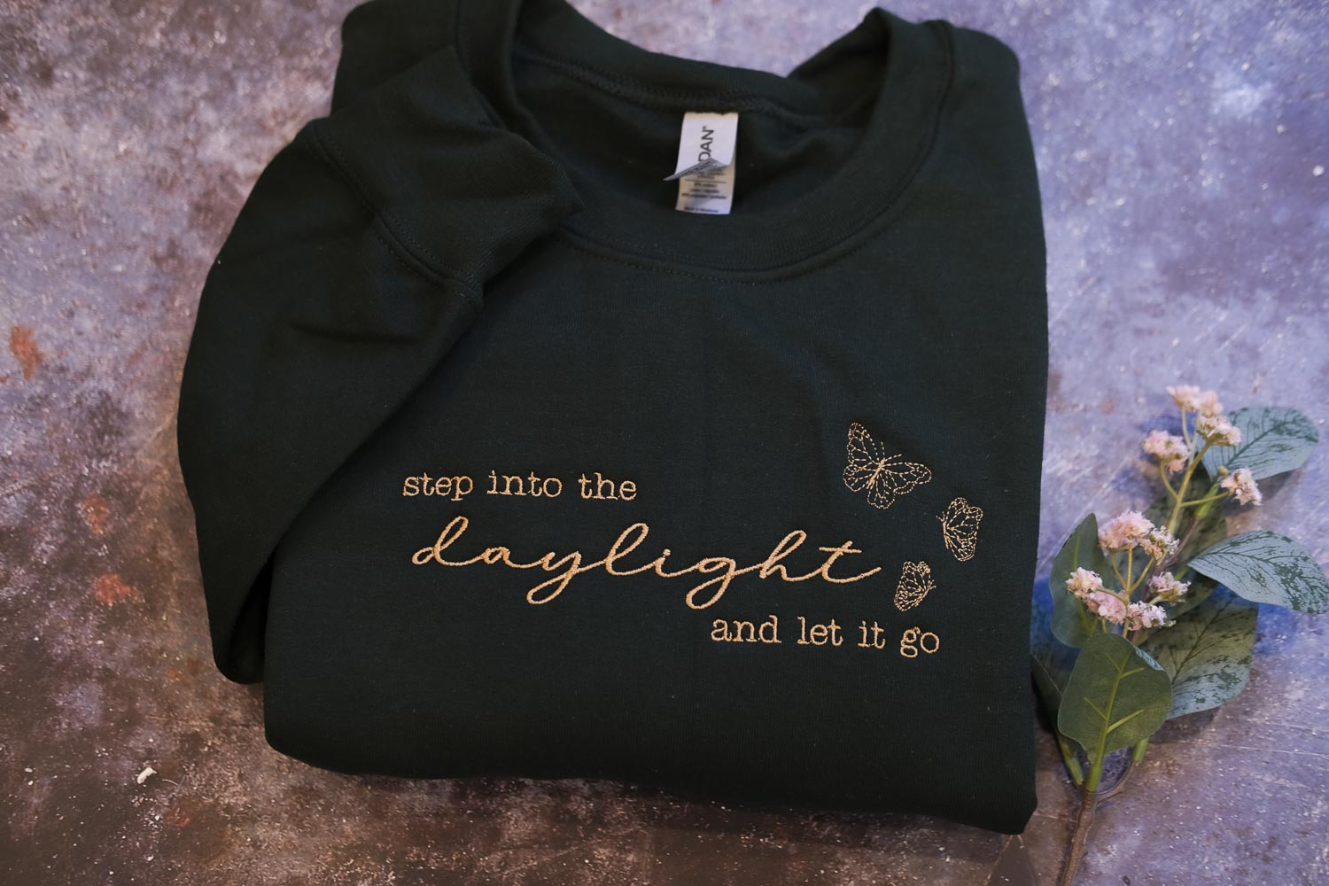 A black sweatshirt with gold text and butterflies printed on it, placed on a textured surface alongside a small plant with white flowers.