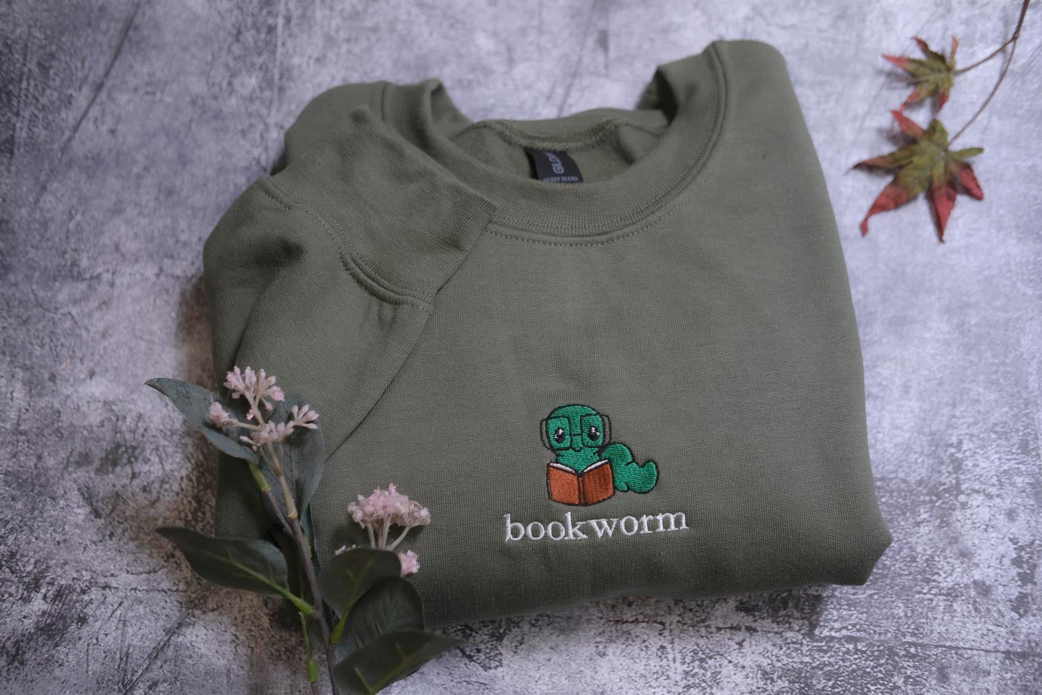 A green sweatshirt with a cartoon image of a caterpillar reading a book, along with the text "bookworm" printed on it, is displayed on a gray surface alongside some pink flowers.