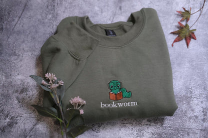 A green sweatshirt with a cartoon image of a caterpillar reading a book, along with the text "bookworm" printed on it, is displayed on a gray surface alongside some pink flowers.