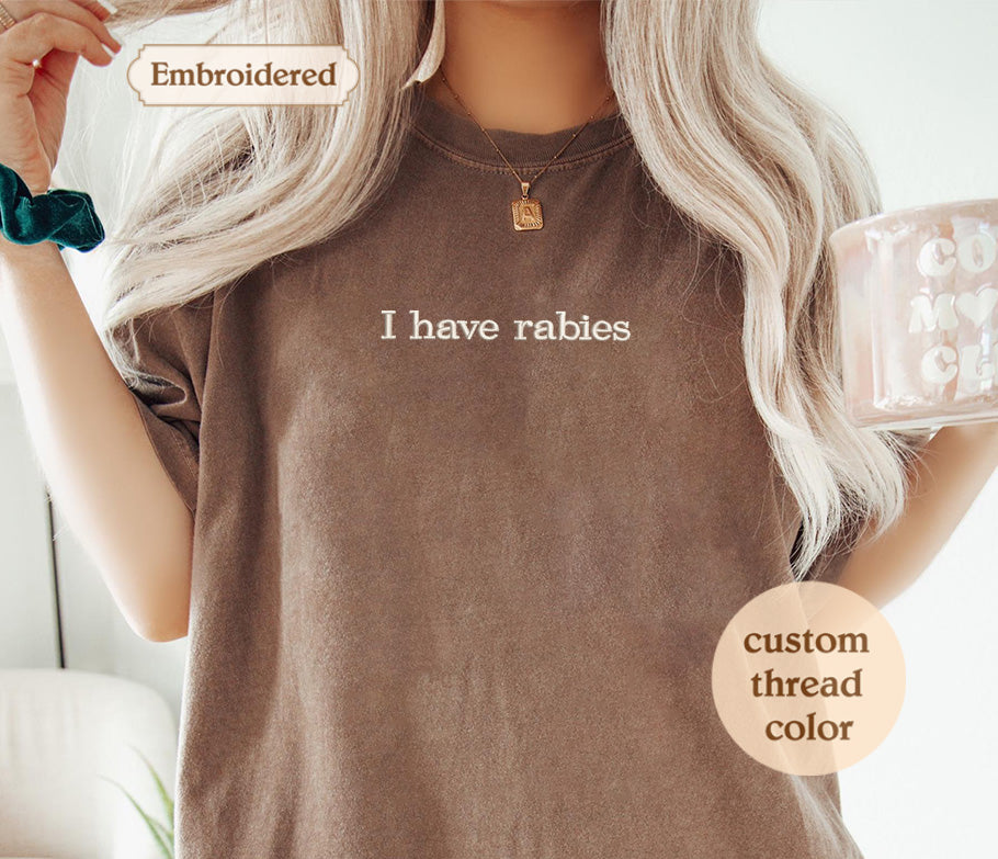 A woman with long blonde hair wearing a brown t-shirt with the text "I haverabies" printed on it. She is holding a mug and has a necklace around her neck.