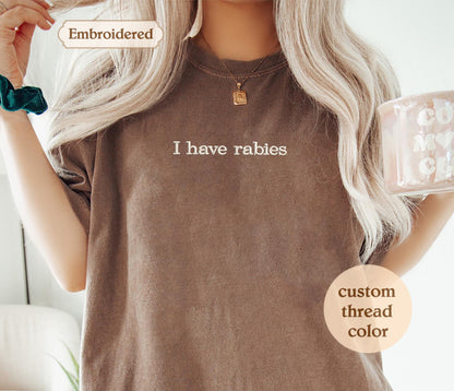 A woman with long blonde hair wearing a brown t-shirt with the text "I haverabies" printed on it. She is holding a mug and has a necklace around her neck.
