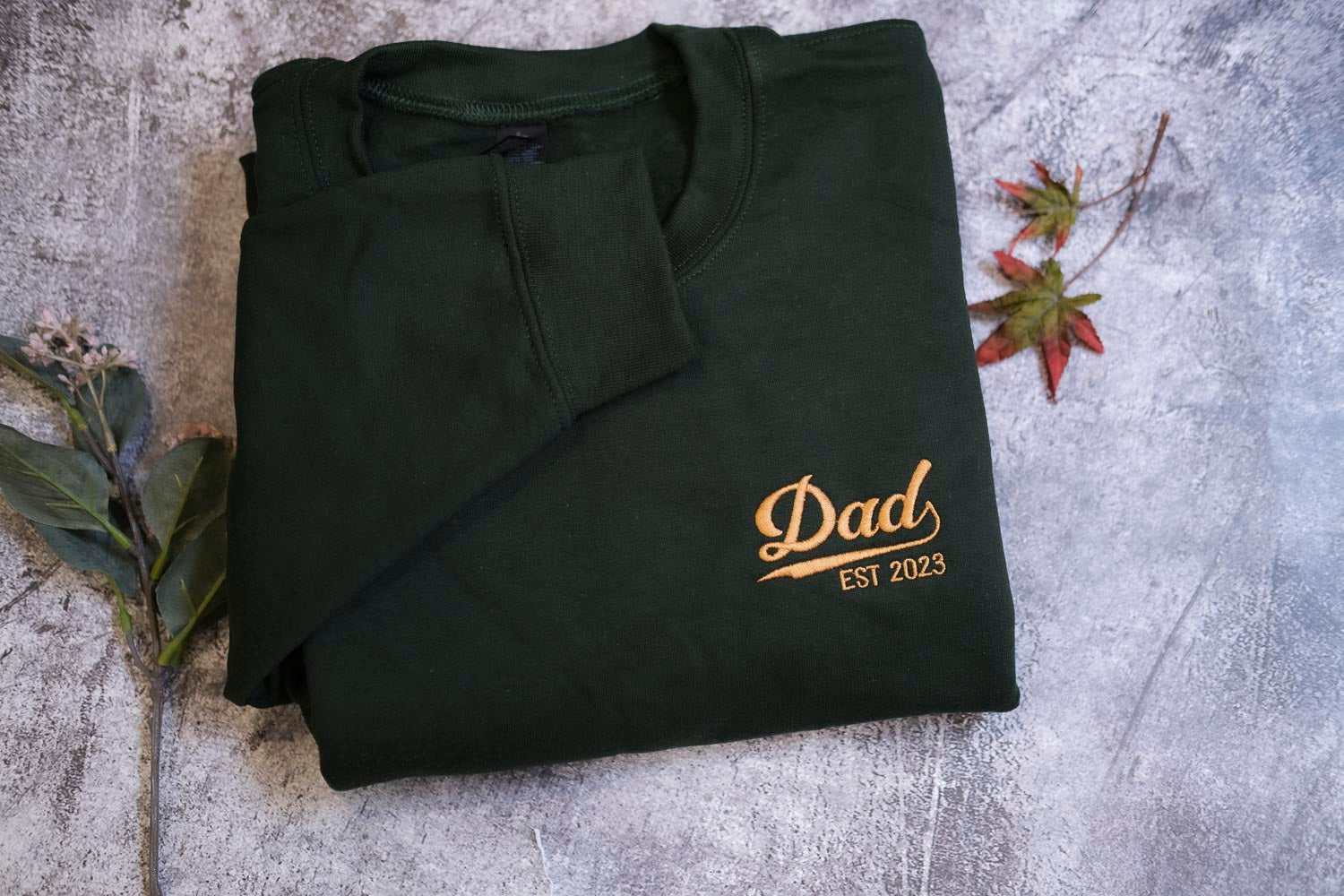 A dark green long-sleeved shirt with the text "Dad EST 2023" printed on it, along with a small leaf and a twig, all placed on a gray surface.