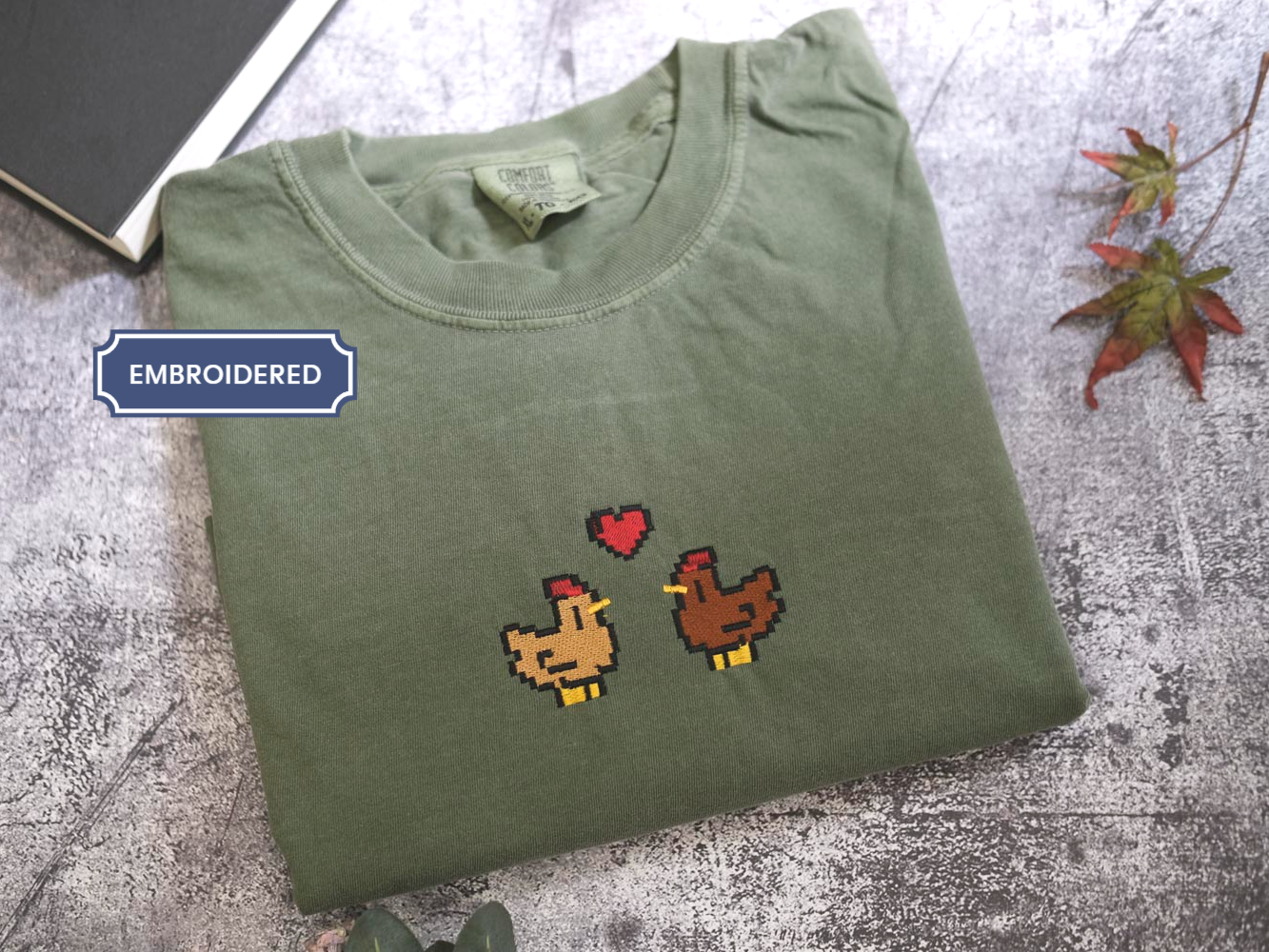 a green t-shirt with a pixelated design of two chickens, one brown and one brown, with a heart between them. The t-shirt is placed on a gray surface, and there is a book and some leaves in the background.