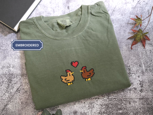 a green t-shirt with a pixelated design of two chickens, one brown and one brown, with a heart between them. The t-shirt is placed on a gray surface, and there is a book and some leaves in the background.