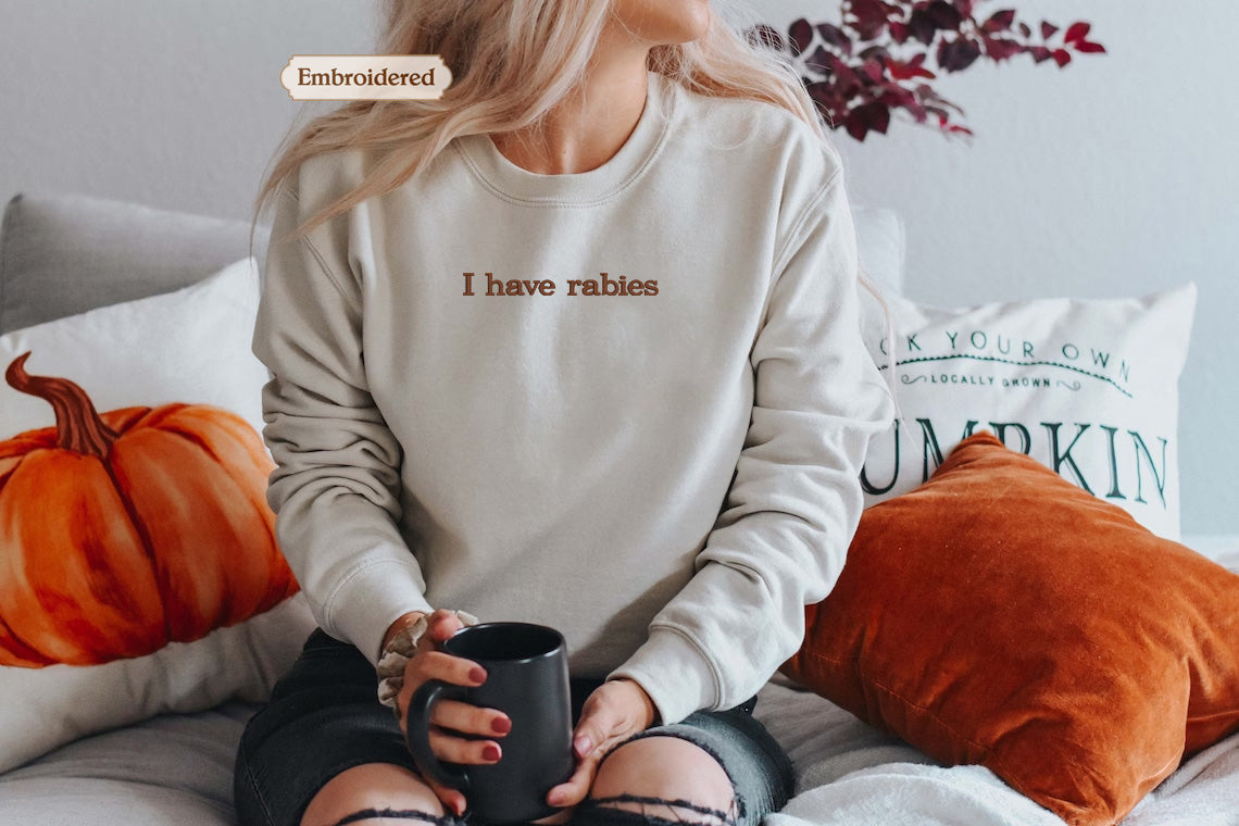 A woman sitting on a bed with a white sweater that says "I have Rabies" embroidered on it. She is holding a black mug and surrounded by pillows with pumpkin designs.