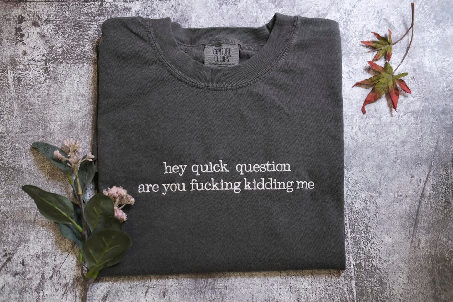 a gray t-shirt with the text "hey quick question are you fucking kidding me" printed on it, along with some leaves and flowers.