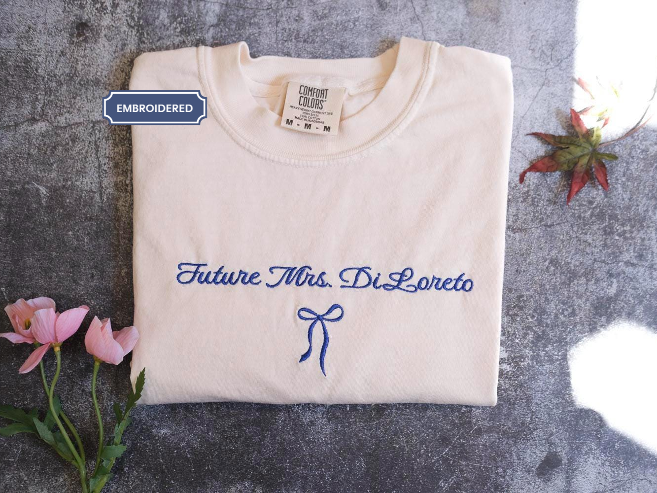a white t-shirt with blue embroidered text and a blue ribbon bow, placed on a gray surface alongside some pink flowers and a small plant.