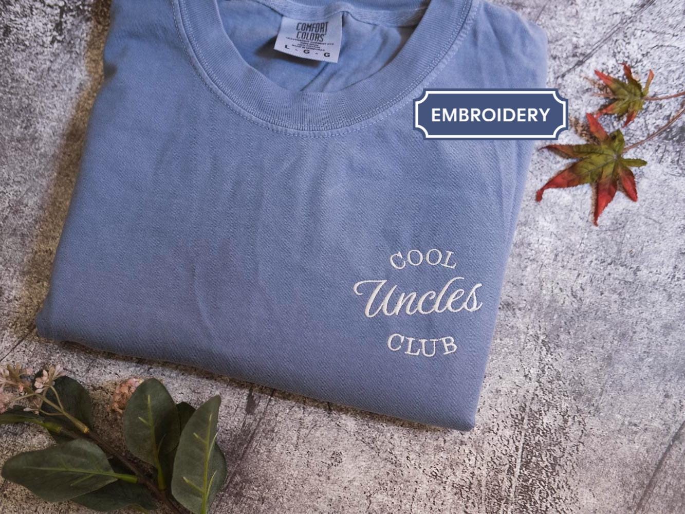 A blue t-shirt with the text &quot;Cool Uncle Club&quot; printed on it, along with some leaves and a small plant.