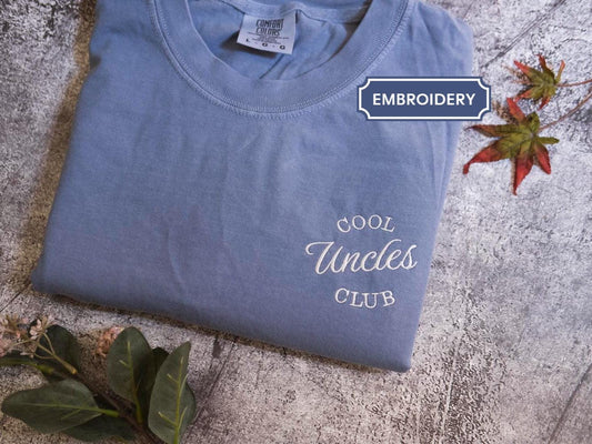 A blue t-shirt with the text &quot;Cool Uncle Club&quot; printed on it, along with some leaves and a small plant.