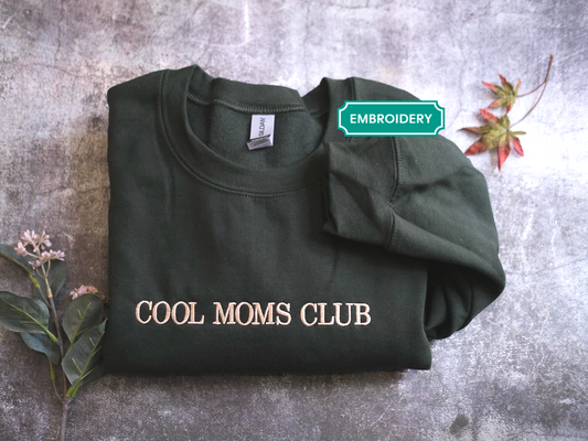 a black t-shirt with the text "COOL MOMS CLUB" printed on it, along with a small plant and a red leaf on the side.