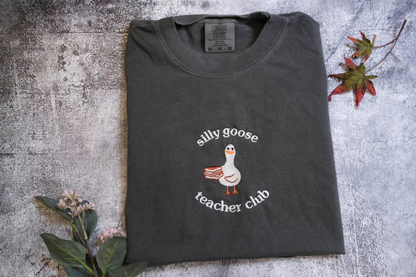 A gray t-shirt with a graphic of a goose and the text "silly goose teacher chub" printed on it, along with a small leaf and a flower.