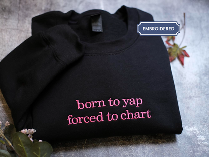 A black sweatshirt with the text "born to yap forced to chart" printed on it, along with a small pink flower and a leaf.