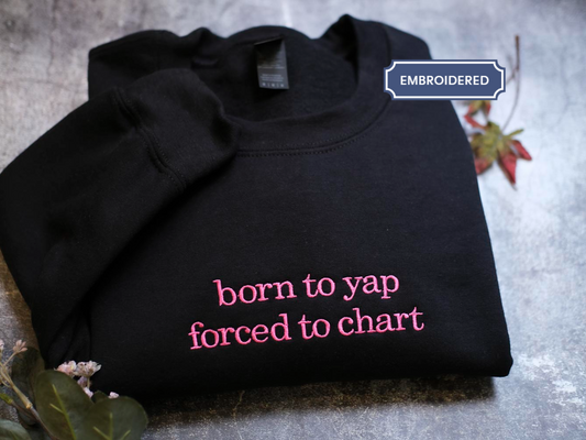 A black sweatshirt with the text "born to yap forced to chart" printed on it, along with a small pink flower and a leaf.