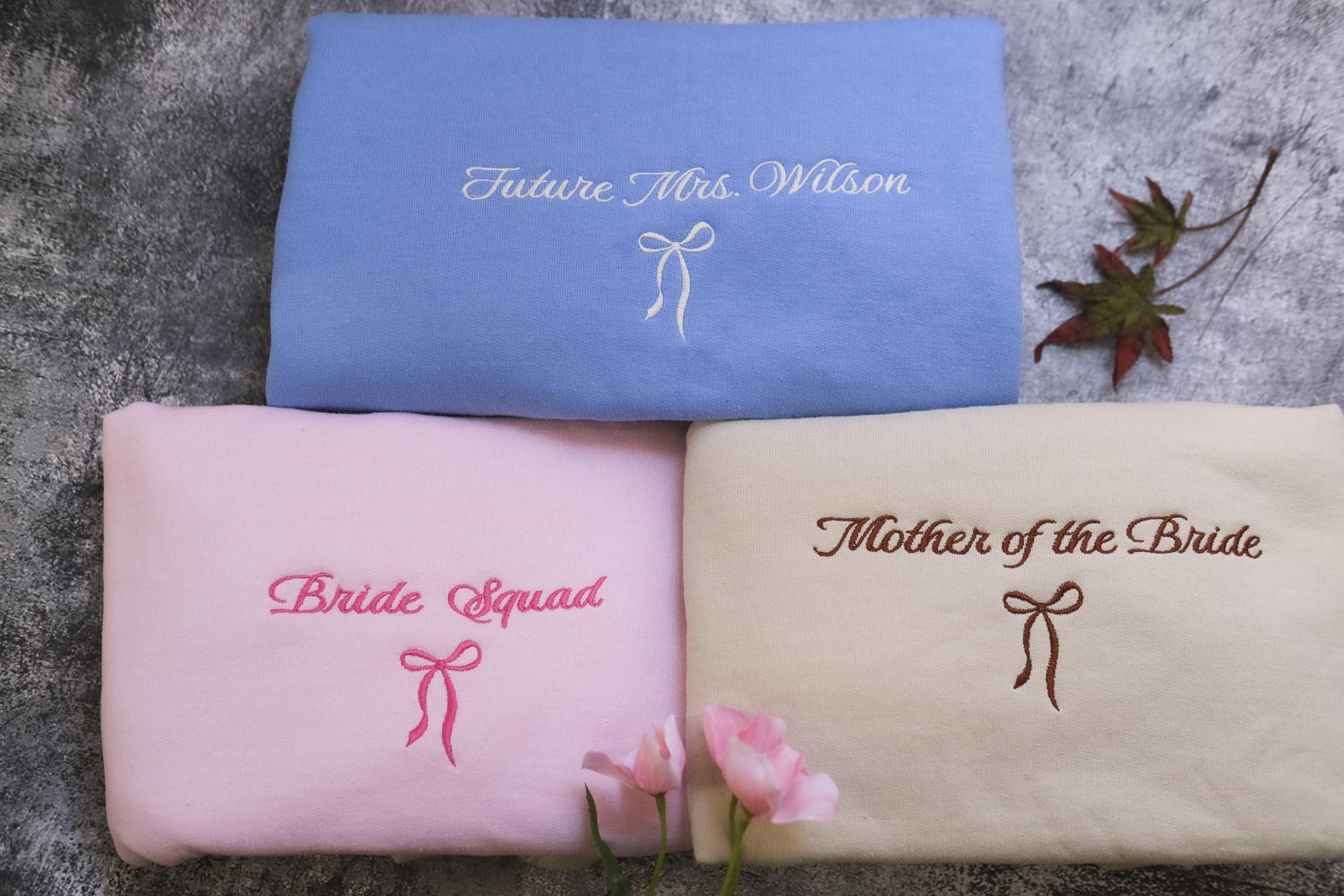 three shirts, each with a different embroidered text and design, arranged on a gray surface.