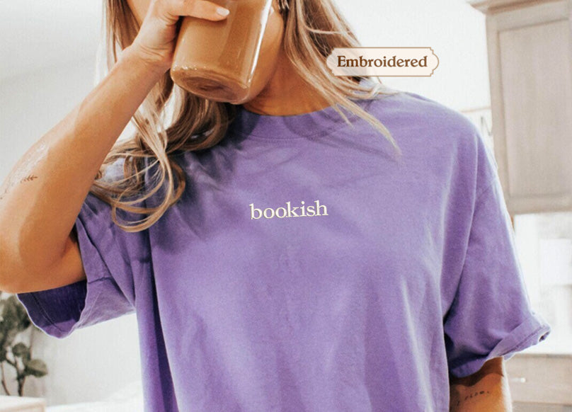 A person wearing a purple t-shirt with the word "bookish" printed on it, holding a drink in their hand.