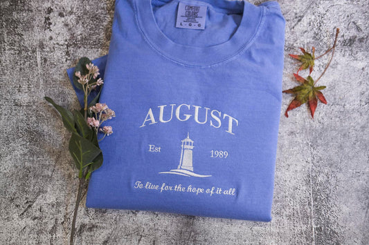 A blue t-shirt with the text "AUGUST Est 1989 To live for the hope of it all" printed on it, along with a lighthouse graphic, is displayed on a textured surface alongside some flowers and a fallen leaf.