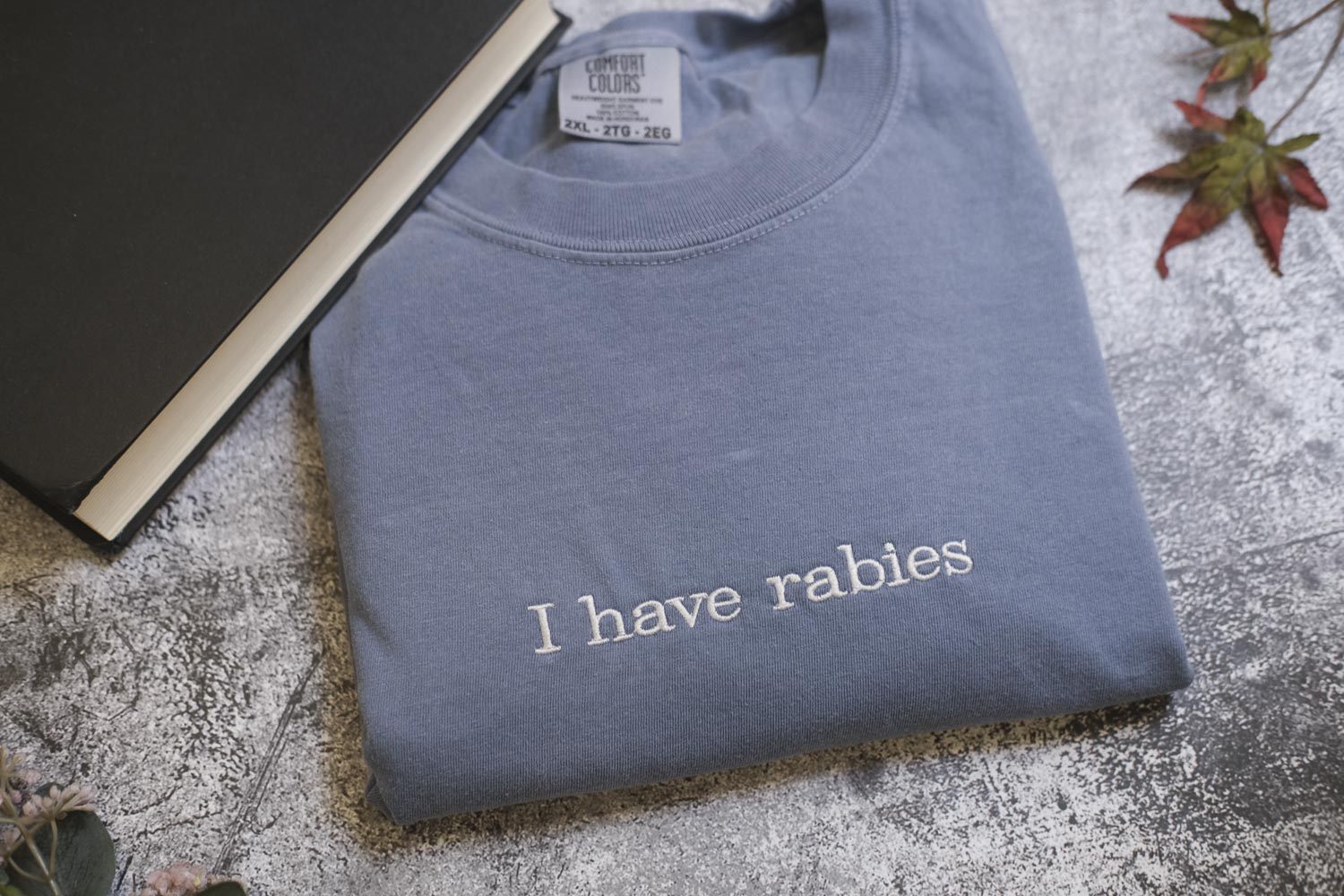 A gray t-shirt with the text "I haverabies" printed on it, along with a black book and some leaves in the background.