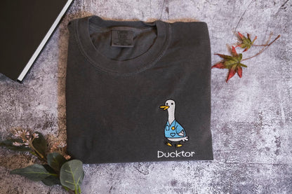 a gray t-shirt with a cartoon duck wearing a blue shirt and the word "Duckster" printed on it, along with a small leaf and a book.
