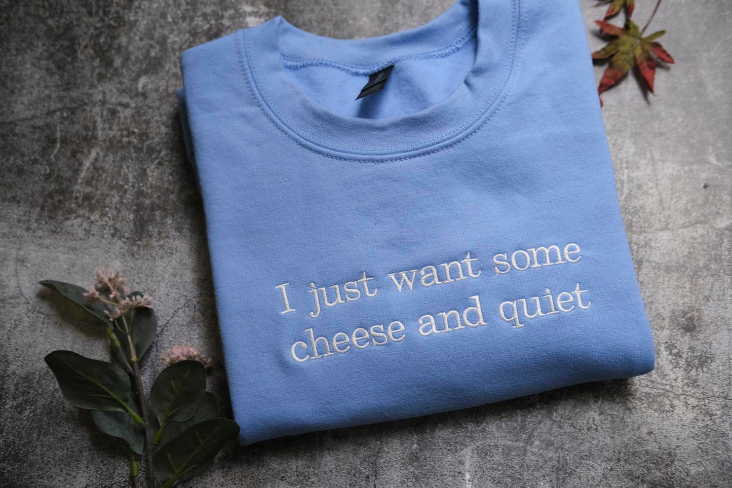 a blue t-shirt with white text that reads "I just want some cheese and quiet". The t-shirt is laid out on a gray surface, with some leaves and flowers nearby.