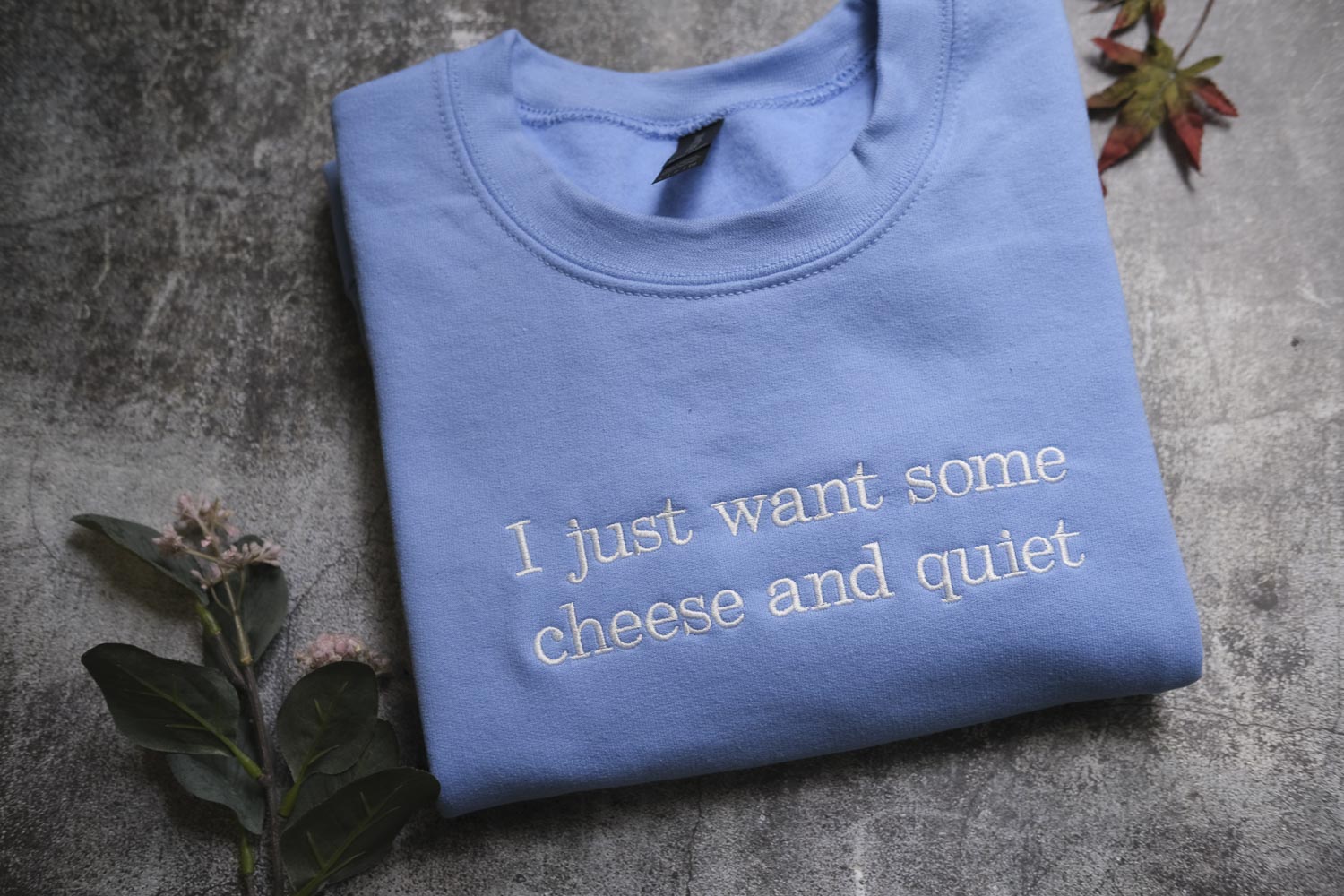 a blue t-shirt with white text that reads "I just want some cheese and quiet". The t-shirt is laid out on a gray surface, with some leaves and flowers nearby.