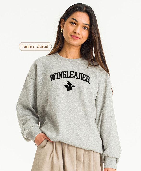 A young woman wearing a grey sweatshirt with the text "WINGLEADER" printed on it, standing against a white background.