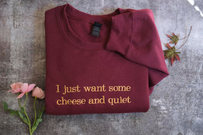 A burgundy sweatshirt with the text "I just want some cheese and quiet" printed on it, along with a small pink rose and some leaves.
