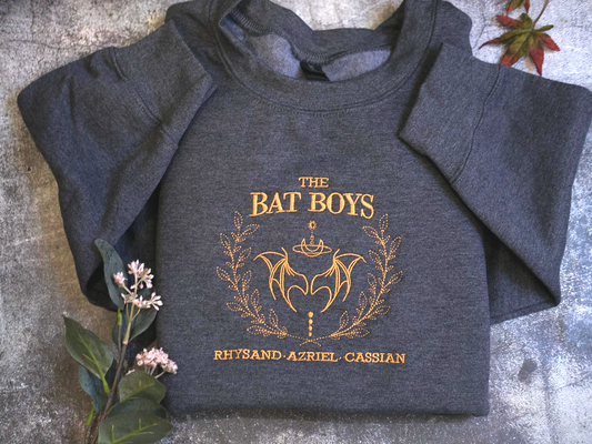 a gray sweatshirt with a gold design and text, placed on a gray surface next to a small plant with pink flowers.