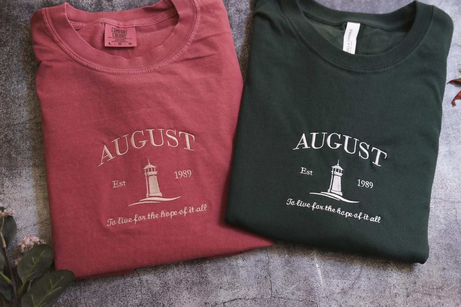 two t-shirts with the word "AUGUST" printed on them, along with the text "Est. 1989" and "To live for the hope of it all" printed below. The t-shirts are laid out on a gray surface, with some leaves visible in the background.