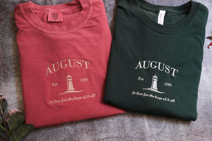 two t-shirts with the word "AUGUST" printed on them, along with the text "Est. 1989" and "To live for the hope of it all" printed below. The t-shirts are laid out on a gray surface, with some leaves visible in the background.