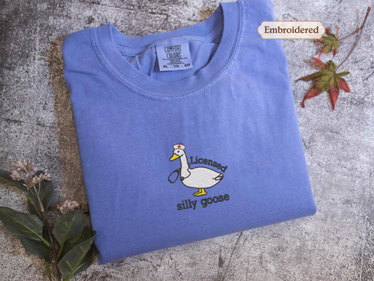 a blue t-shirt with a cartoon goose and the text "Licensed silly goose" embroidered on it. The t-shirt is placed on a gray surface, with some leaves and flowers visible in the background.