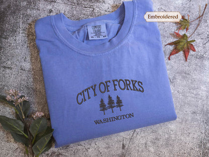 a blue t-shirt with the text "CITY OF FORKS WASHINGTON" printed on it, along with a small embroidered label. The t-shirt is laid out on a textured surface, and there are some leaves and flowers nearby.