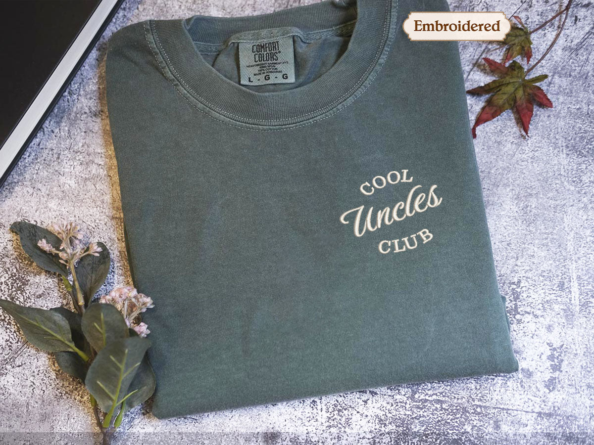 a gray t-shirt with the text "Cool Uncle Club" embroidered on it, placed on a gray surface with some leaves and flowers.