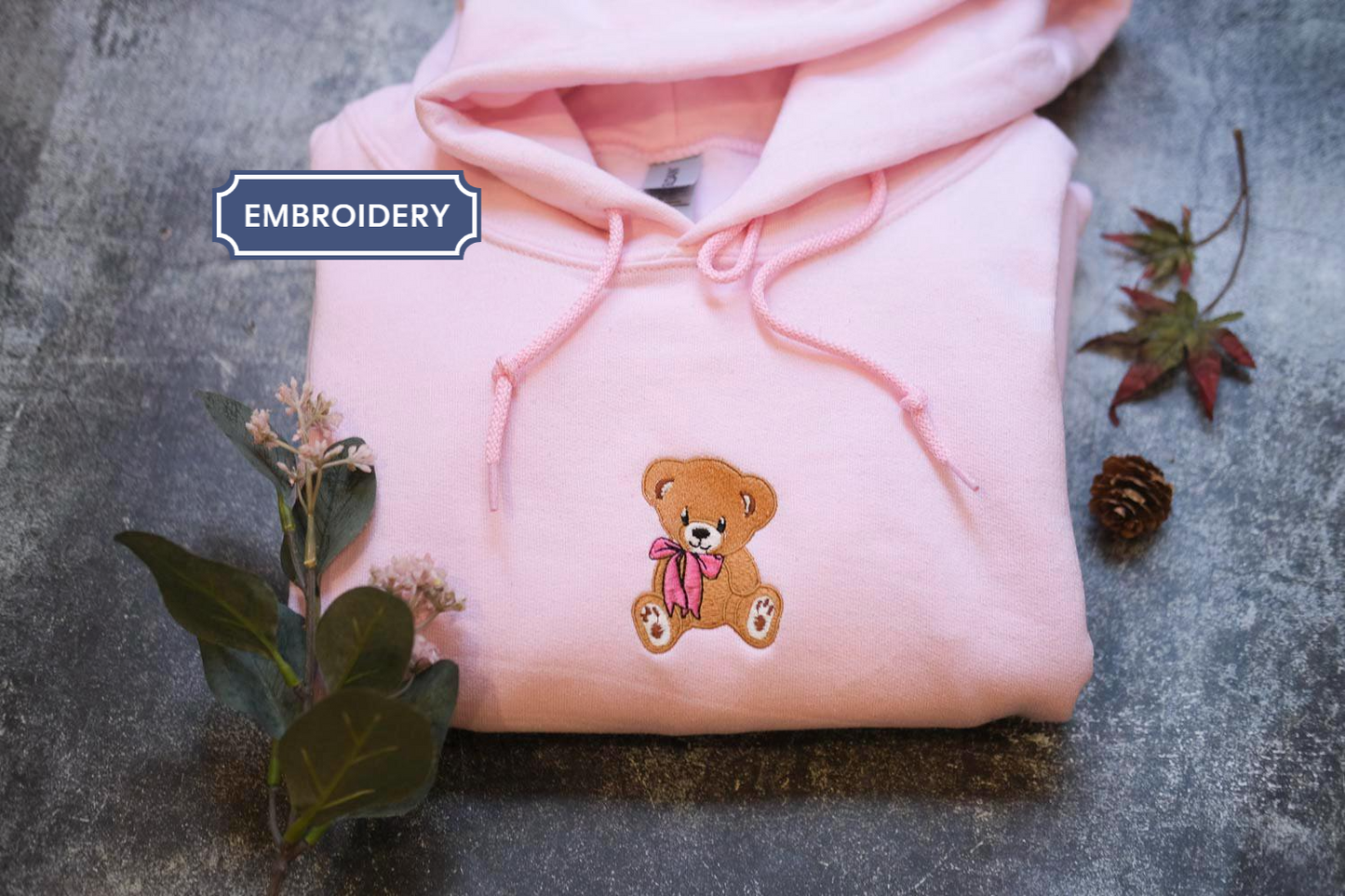A pink hoodie with a teddy bear embroidered on it, along with some flowers and a pine cone, is displayed on a gray surface.