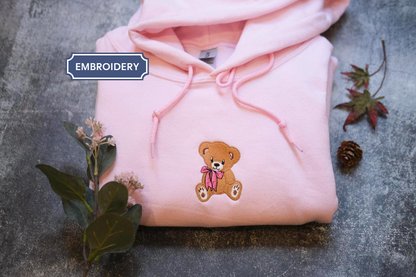 A pink hoodie with a teddy bear embroidered on it, along with some flowers and a pine cone, is displayed on a gray surface.