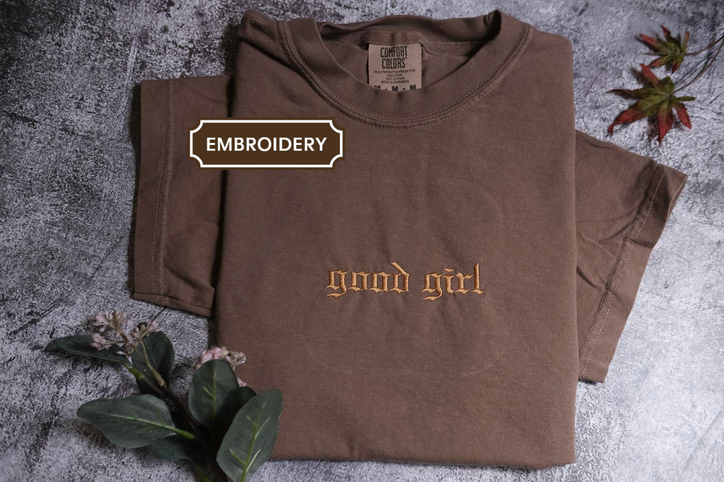 a brown t-shirt with the text "Good girl" printed on it, along with some leaves and flowers on a textured surface.