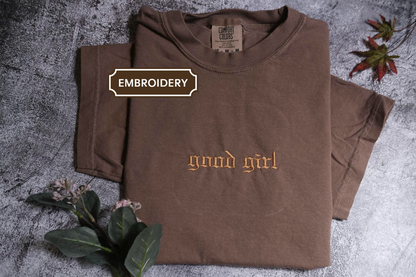 a brown t-shirt with the text "Good girl" printed on it, along with some leaves and flowers on a textured surface.