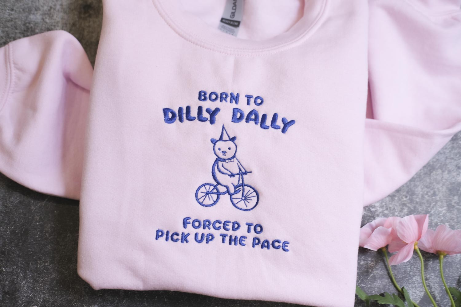 A pink t-shirt with a blue embroidered design of a cat riding a bicycle, along with the text "BORN TO DILLY DALLY FORCED TO PICK UP THE PACE".