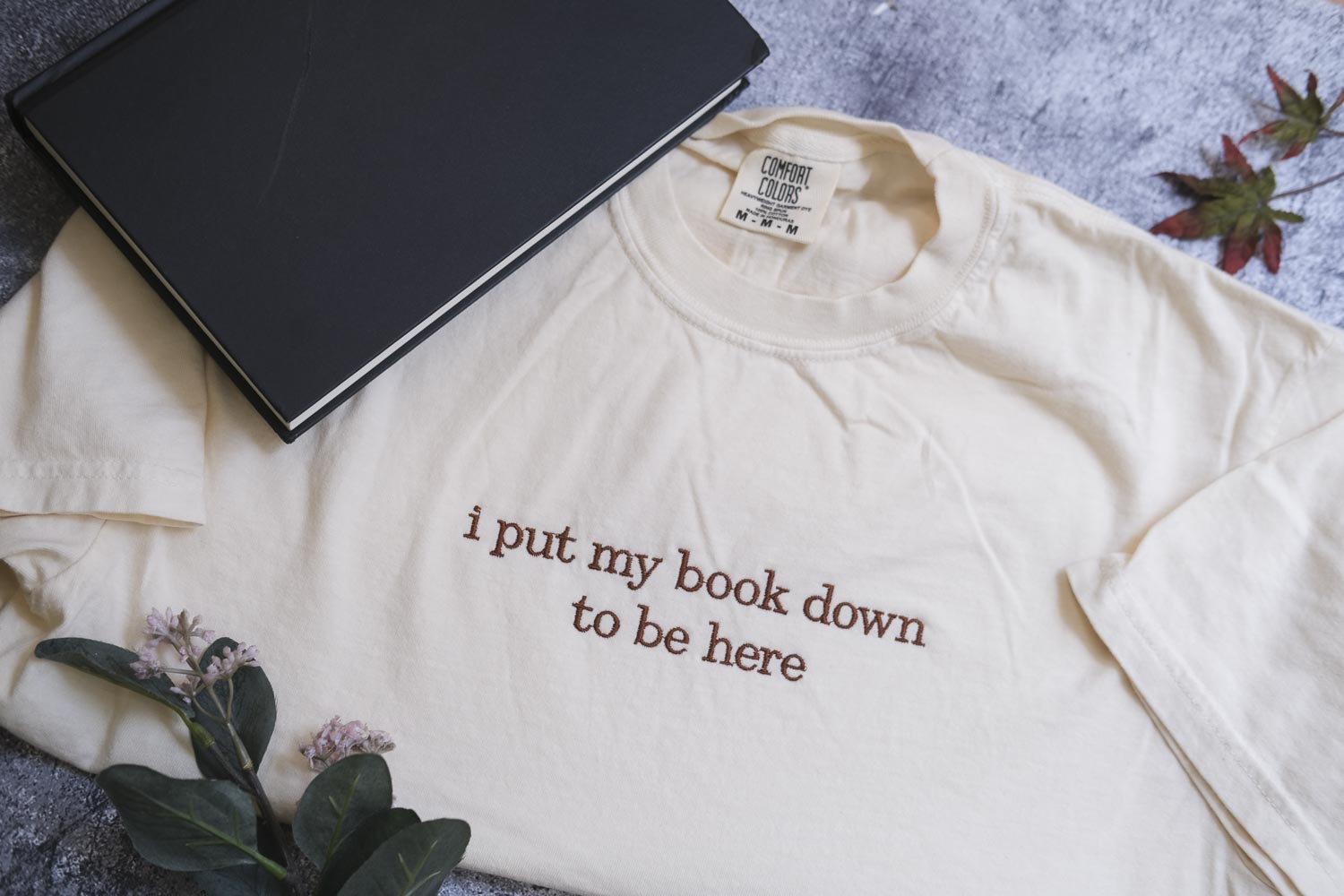 a white t-shirt with the text "I put my book down to be here" printed on it, along with a black notebook and some leaves.