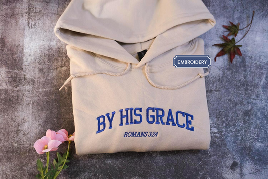 a beige hoodie with blue text and embroidery, placed on a gray surface alongside a small pink flower and a leaf.