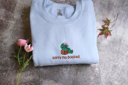 A light blue t-shirt with a cartoon character and the text "Sorry I'm booked" printed on it, along with a small pink flower and a fallen leaf.