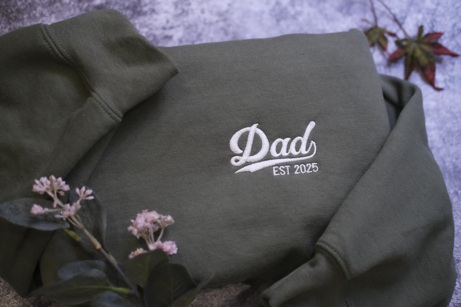 A dark green sweatshirt with the text "DAD EST 2025" embroidered on it, along with some pink flowers and a leaf in the background.