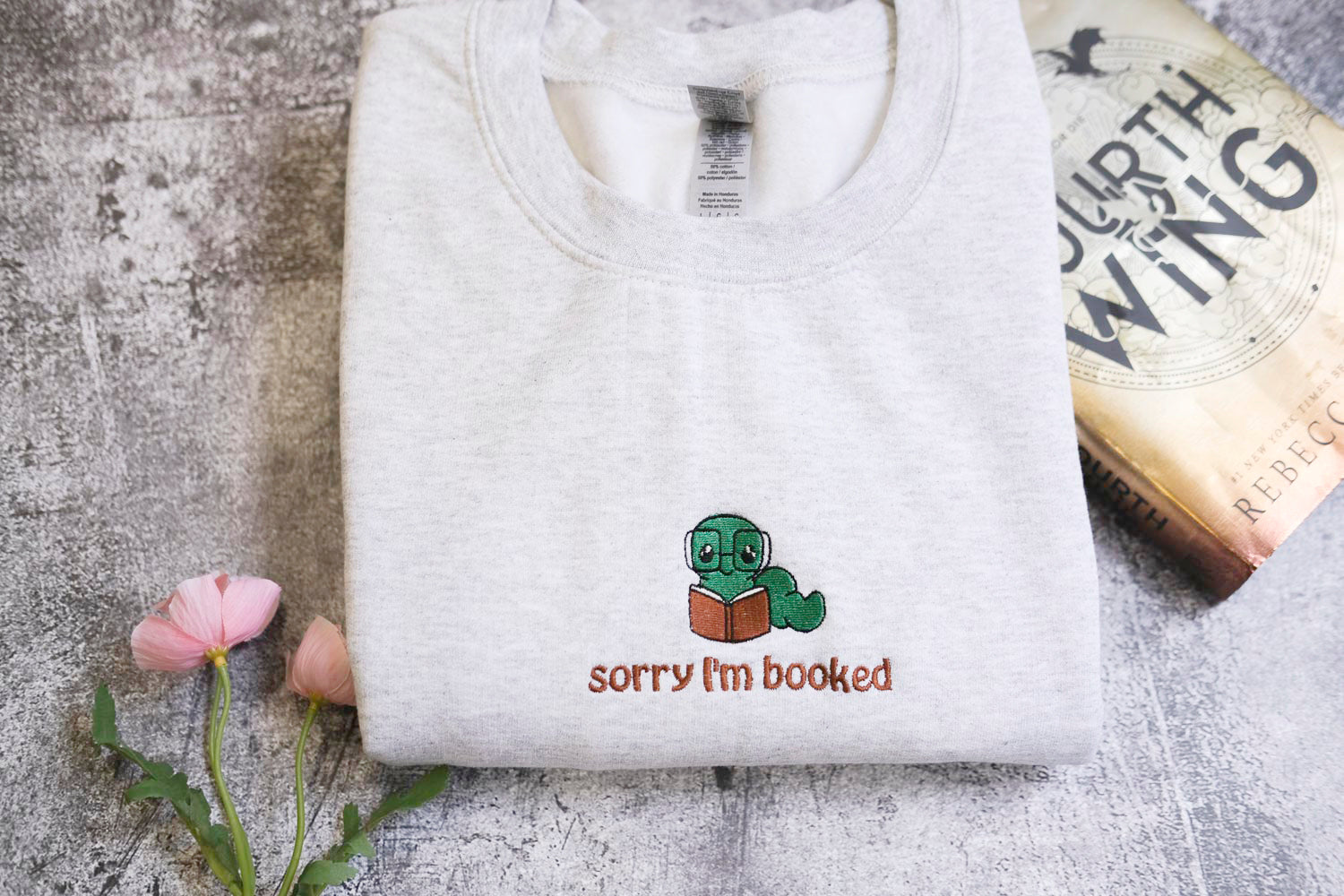 a white sweatshirt with a small embroidered character reading a book, accompanied by a book titled "Fourth of July" and a pink flower.