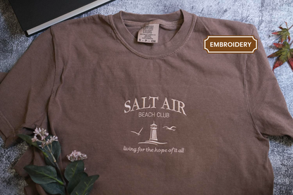 A brown t-shirt with the text "SALT AIR BEACH CLUB" printed on it, along with a lighthouse and birds, is displayed on a gray surface.
