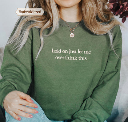 A person wearing a green sweater with the text "hold on just let me overthink this" embroidered on it.