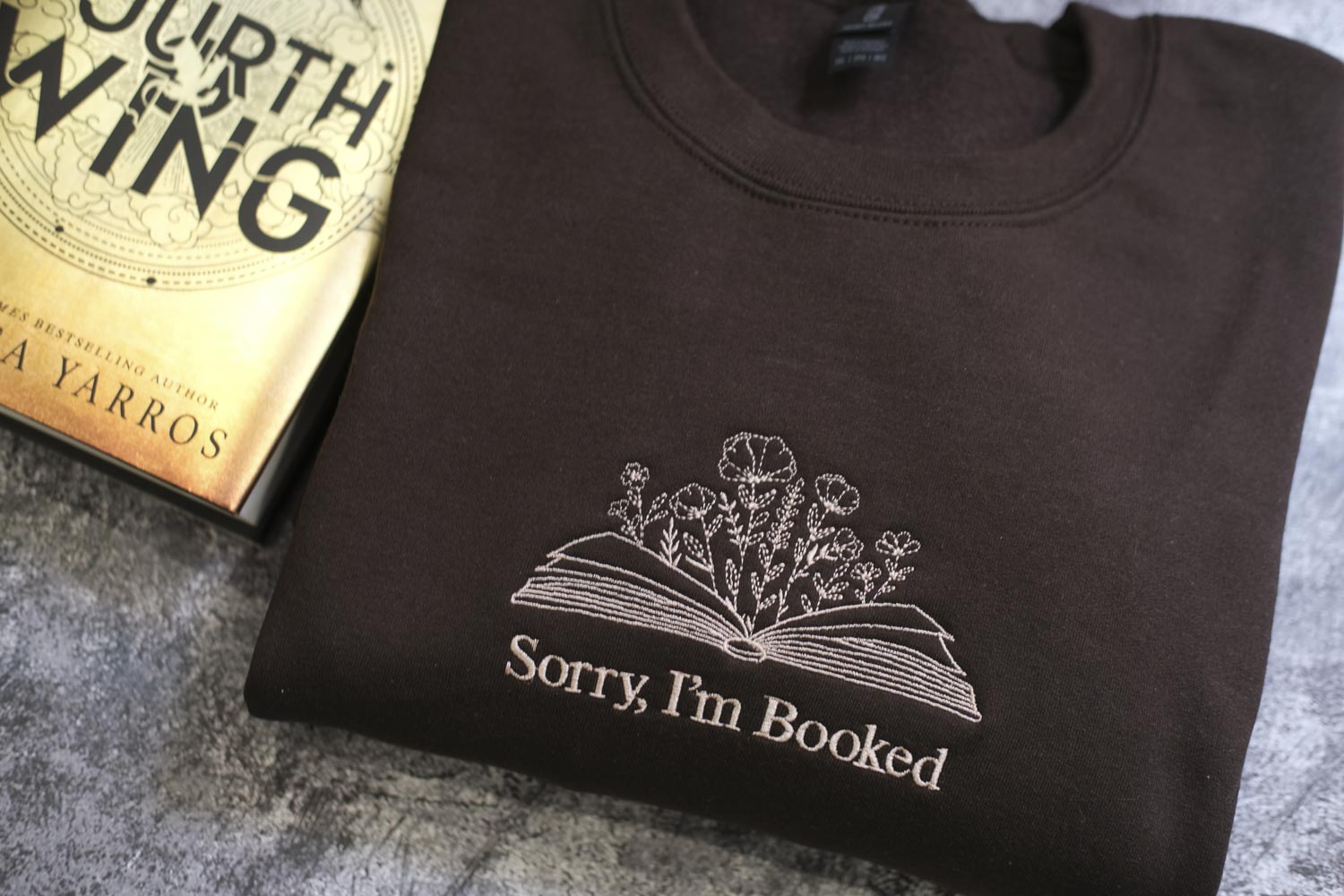A dark-colored sweatshirt with a white embroidered design on the front, featuring a book and flowers, and the text "Sorry, I'm Booked" printed on it.
