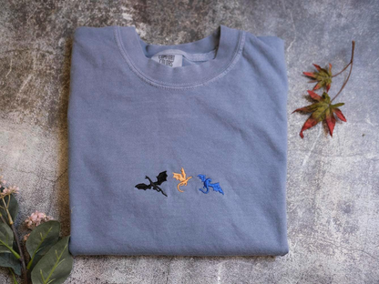 a gray t-shirt with three dragon embroidered designs on it, placed on a gray surface alongside some leaves.