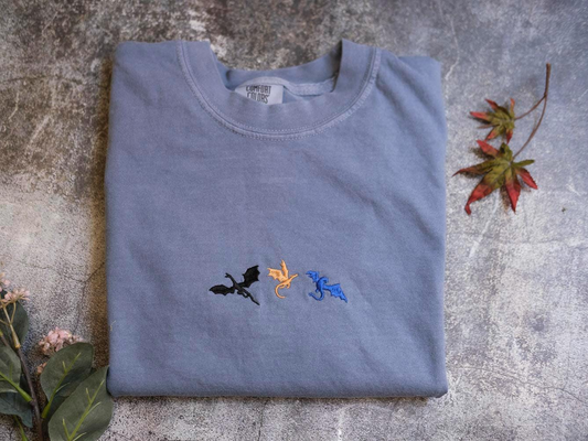 a gray t-shirt with three dragon embroidered designs on it, placed on a gray surface alongside some leaves.