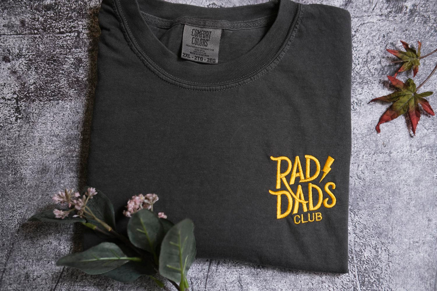 a black t-shirt with yellow text and graphics, including the words "RAD DAD'S CLUB" and a graphic design, placed on a gray surface with some flowers and a leaf.
