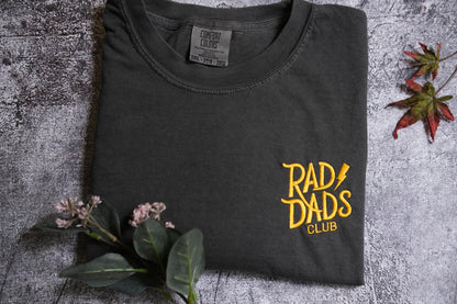 a black t-shirt with yellow text and graphics, including the words "RAD DAD'S CLUB" and a graphic design, placed on a gray surface with some flowers and a leaf.