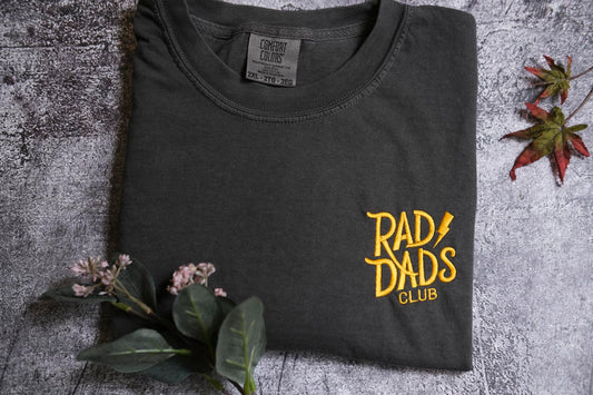 a black t-shirt with yellow text and graphics, including the words "RAD DAD'S CLUB" and a graphic design, placed on a gray surface with some flowers and a leaf.